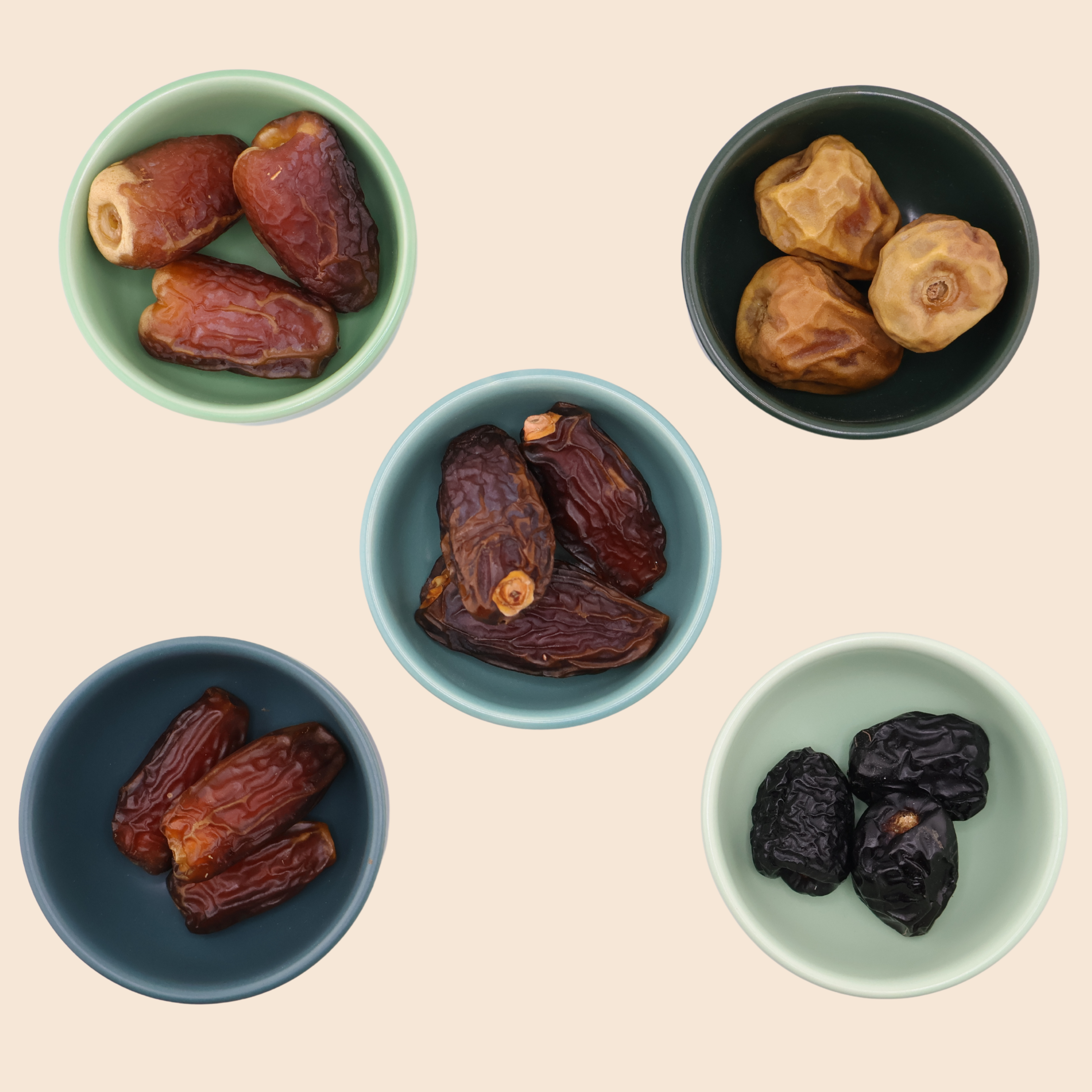 Five small bowls displaying Mighty Mejdool date varieties: Sukkari, Ajwa, Mabroom, Sagai, and Medjool. On a light beige background