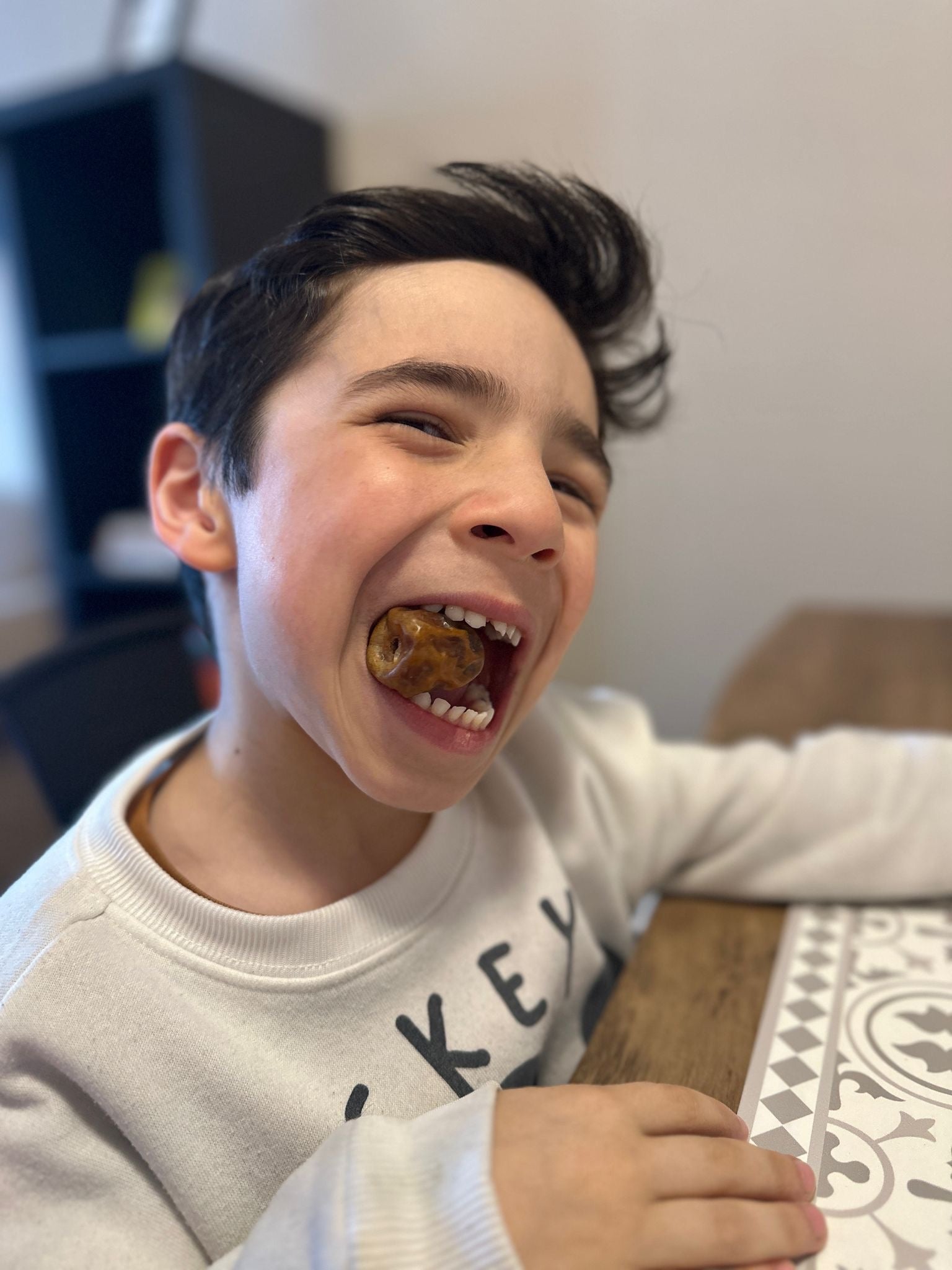 A young boy smiling while eating a Mighty Mejdool date at the table, enjoying a naturally sweet and healthy snack
